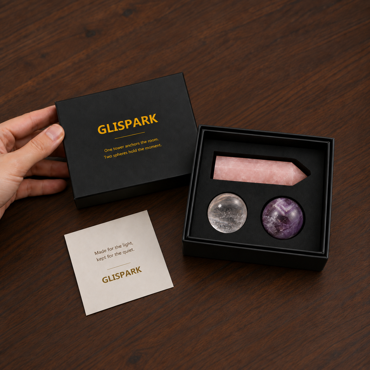 A hand holds a black GLISPARK gift box beside the open Soft Dream Crystal Set with a rose quartz tower, clear quartz sphere, dream amethyst sphere, and brand card on a dark wood table.